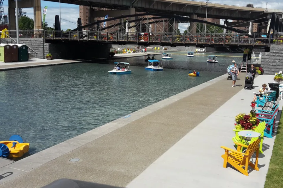 A canal under a bridge in Buffalo is lined by chairs and tables, people walking about, looking at peddle boats on the water.